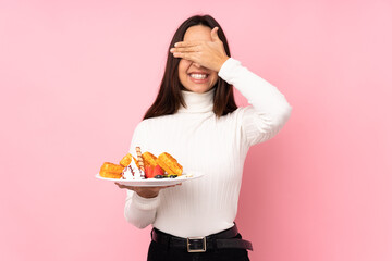 Young brunette woman holding waffles over isolated pink background covering eyes by hands