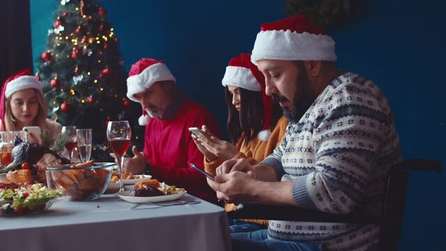 Family with man in wheelchair uses phones during Christmas holiday. Group of friends use their smartphones at festive dinner table. Dependence on phone. Nomophobia