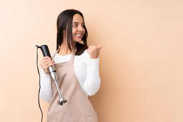 Young brunette girl using hand blender over isolated background pointing to the side to present a product