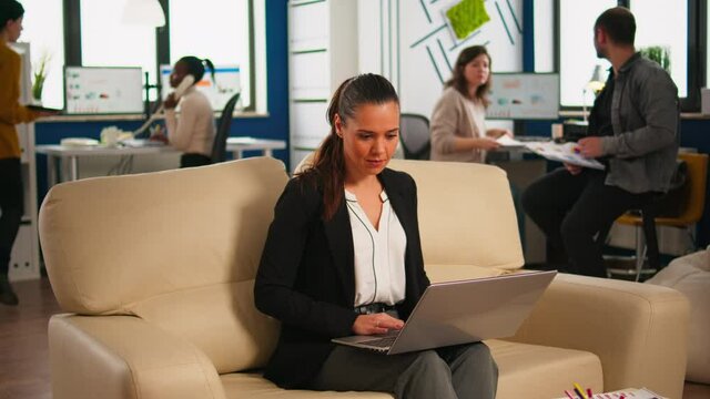 Happy hispanic lady sitting on couch in start up business office typing on laptop while diverse colleagues working in background. Multiethnic coworkers planning new financial project in modern company