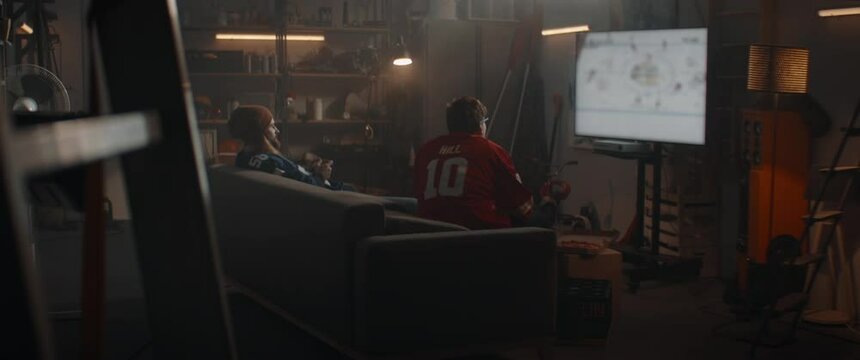 Two friends playing hockey sport video game inside garage hideout, enjoying pizza and drinks. TV screen is blurred. Shot with 2x anamorphic lens