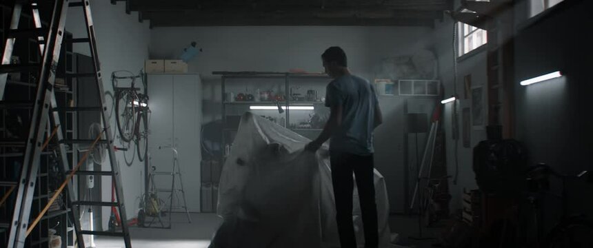 Caucasian male biker taking off cover from classic vintage motorcycle inside garage. Shot with 2x anamorphic lens