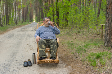 Senior man drink cup of tea while resting in spring  forest sitting on a wicker rocking-chair.