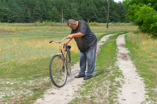 Senior Ukrainian peasant standing on a country road and inspecting old rusty bicycle with broken saddle