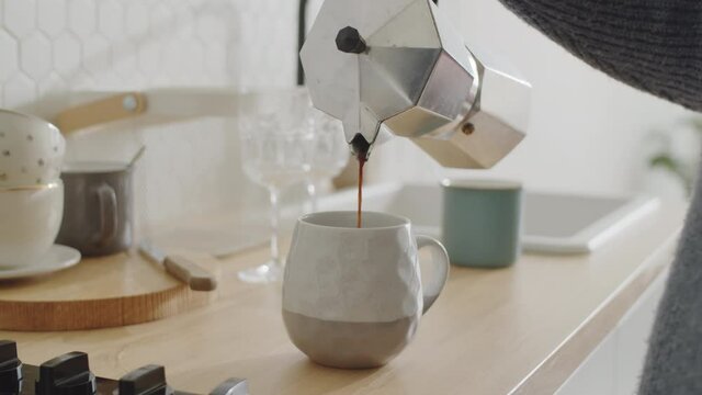 Close up shot of hand of unrecognizable woman pouring coffee into cup from steel espresso maker while preparing morning drink in the kitchen at home