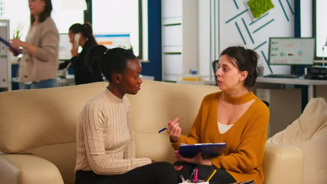 Diverse businesswomen discussing new project for company evolution, black woman checking tasks on clipboard, signing new contract. Multiethnic employees gathered in co-working space
