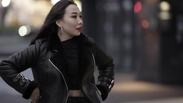 Portrait of a beautiful and spectacular Asian woman with long dark hair with red lipstick on her lips in a stylish image, she is poses on a city street against the background of evening lights.