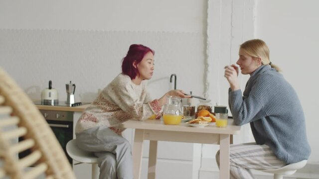 Young Asian woman discussing social media on smartphone with Caucasian female flatmate while having breakfast at kitchen table in the morning at home