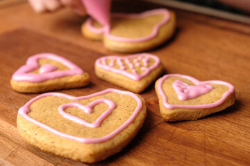 Icing of Valentines Day. Woman decorating gingerbread cookies in the shape of heart on a wooden table.