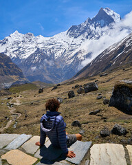 Naklejka premium Hiker sitting on a rock in an alpine meadow gazing at the snow-capped Machapuchare peak at Annapurna Base Camp, Nepal Himalayas
