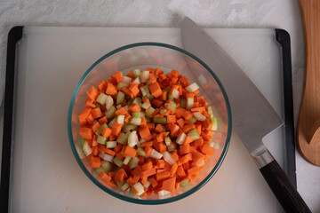 Carrots, Onions and Celery being Sliced on White Cutting Board With Chef's Knife and Wood Stand in a Bowl