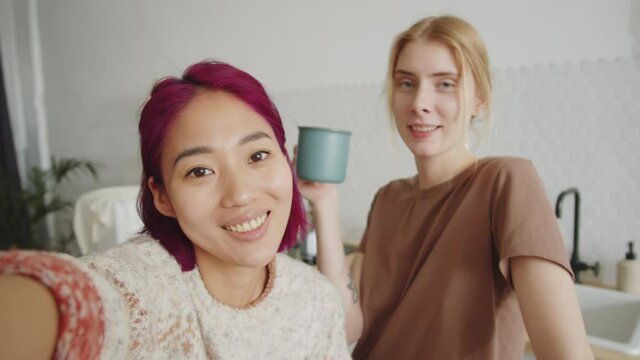 Young cheerful Asian and Caucasian female friends waving, smiling and chatting at camera while video calling from home in the morning