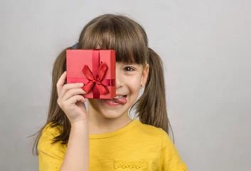 Gordijnen Foto van een aantrekkelijk klein meisje met lange rechte paardenstaarten houdt in een hand een rode geschenkdoos met strik. Gelukkig meisje met cadeau toont haar tong en glimlacht © Albert Ziganshin