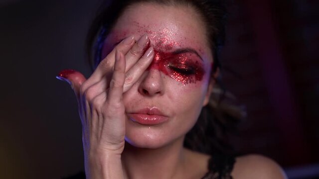 Portrait of an adult dark-haired woman applies unusual makeup, paints around the eyes with red lipstick. The top covers everything with red sequins.