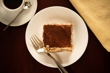 Slice of Traditional Italian Tiramisu on White Plate and Wooden Table with Fork and Coffee Cup.