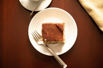 Slice of Traditional Italian Tiramisu on White Plate and Wooden Table with Fork and Coffee Cup.