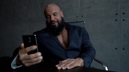 muscular Jock a bald man with a beard sits at a table and uses a mobile phone, talks on a video link and smiles.