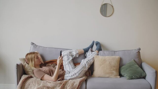 Side view of young blonde woman in comfortable homewear lying on sofa with legs up and using smartphone