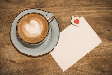 Beautiful cup of coffee with a heart shape and white blank sheet of paper on a wooden background. Valentine's day concept. Selective focus.
