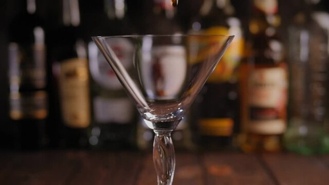 Close-up of a pink Martini being poured into a Martini glass in a bar against the background of a blurred bar counter with various bottles of alcohol, slow motion.