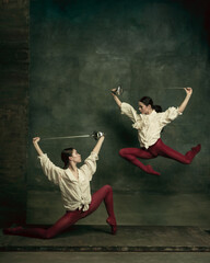 Naklejka na ściany i meble Flying. Two young female ballet dancers like duelists with swords on dark green background. Caucasian models dancing together. Ballet and contemporary choreography concept. Creative art photo.