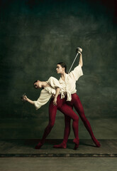 Passioned. Two young female ballet dancers like duelists with swords on dark green background. Caucasian models dancing together. Ballet and contemporary choreography concept. Creative art photo.