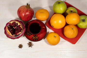 Tangerines, apples and an orange are on the red platter. Nearby is a cup of juice and pomegranate and star anise.