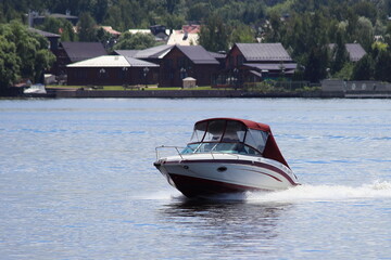 Fototapeta premium Fast gliding white motor boat front side view on cottage houses and green trees on far shore background in Moscow area at Sunny summer day, countryside active boating tourism recreation on river water