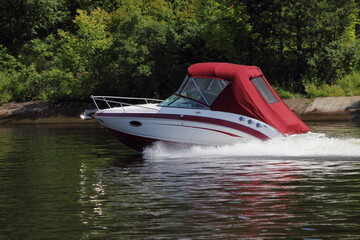Fototapeta premium Fast gliding red tented inboard motor boat side view on green trees on far shore background in Moscow area at Sunny summer day, active boating tourism recreation on river