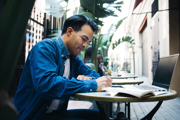 Smiling ethnic distance worker writing on paper near laptop outdoors