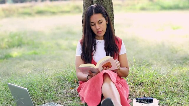 Acercamiento a rostro de Mujer latina leyendo un libro en el parque, sentada en el c&eacute;sped recargada de un &aacute;rbol.	