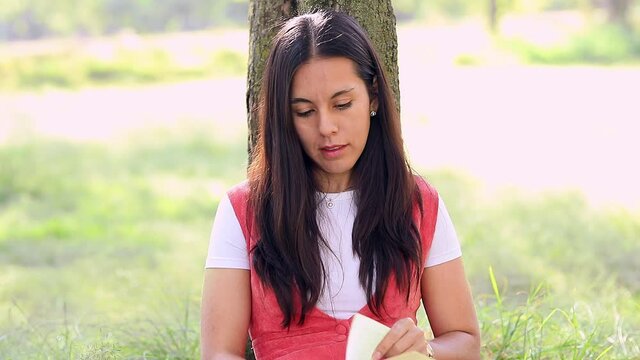 Acercamiento a rostro de Mujer latina leyendo un libro en el parque, sentada en el c&eacute;sped recargada de un &aacute;rbol.	