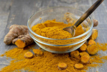 Fresh turmeric root and turmeric powder in a transparent bowl on a gray background.