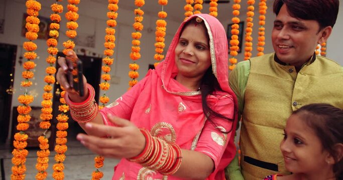 Young couple with daughter wearing colorful traditional dress taking photos selfie on smart phone mobile technology device camera digital on Hindu festival of Diwali, day outdoors courtyard