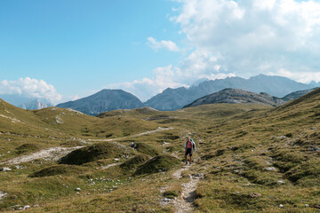Fototapeta premium A woman hiking along a high plateau in Italian Dolomites. High mountains around. The area is partially overgrown with grass and partially covered with stones. Remote and desolate area. Freedom