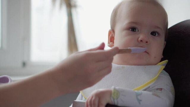 Food, Nutrition, Childhood Concepts - Mom Feeds Infant Baby With Rubber Spoon. Cute Not Hungry Child Dressed In Bib Sits In Kids Chair Dont Want To Eat Dish In Kitchen Indoors. Dislike First Feeding