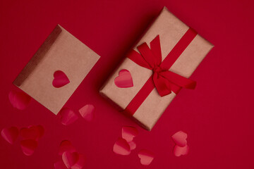 Valentine day composition with copy space. Present or gift box wraped in craft paper with ribbon bow and red hearts. Envelope postcard with confetti on red background. Close up, top view, copy space