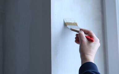 a man's hand with a small bright red brush applies a layer of white acrylic paint to the surface of...