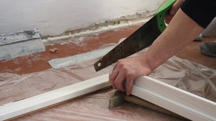 male hands of a carpenter working with a hand saw in preparation for installing a door frame made of light material against the background of the floor and old walls