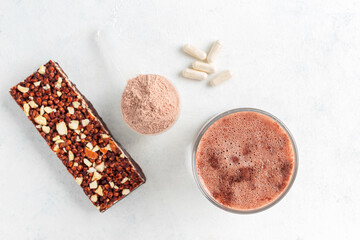 Glass with milkshake of whey protein isolate, protein powder in scoop, granola bar, white capsules of amino acids and creatine, bodybuilding food supplements on white background, top view, flat lay
