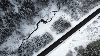 Luftaufnahme mit einer Drohne von einer Straße mit einem Auto das im Winter durch den Wald fährt
