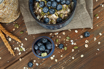 Oatmeal porridge with blueberries and seeds in a blue ceramic bowl and a glass jar of oat flakes on a wooden background. Healthy eating concept. Top view.