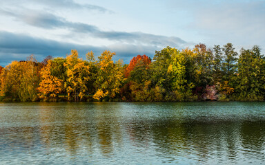 Autumn landscape at lake, multicolored trees, water mirror, sky clouds in background
