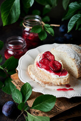 Atmospheric close-up shot of plum jam on a cream cheese toast. Freshly baked bread with few slices cut of it, on a vintage plate, surrounded by plum branches and plums, fresh plum jam in glass jars