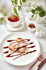 Breakfast table with tea. Cheese pancakes, or syrniki, on a white plate are styled with chocolate sauce. Blooming bird cherry in a vase as table decoration. Light fresh springtime atmosphere