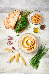 Freshly made hummus with chickpeas, olive oil, paprika in glass bowl on light grey table, ingredients freely arranged around. Flatbreads and fresh parsley to go with hummus on a small chopping board