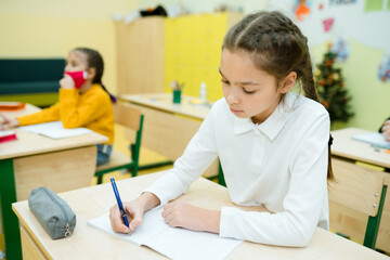 Little girl in a white shirt with gathered hair sits at a desk and writes in a notebook
