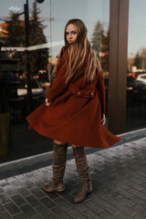 Fototapeta premium young blonde woman in autumn clothes walks on street background shop windows. Street style Fashion 2021 Winter Spring Terracotta Coat Woolen Cashmere Suede Over Knee Boots White Golf