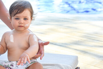 Mother applying body cream or sunscreen on her cute baby near swimming pool. Summer vacation with children. Family holiday on resort.  Mother love, mother care and happy motherhood concept. Healthcare
