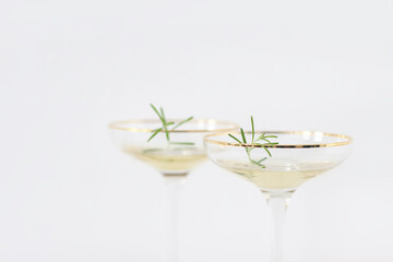 Happy New Year still life. Close-up of two champagne wine glasses with golden rim and green rosemary leaves. Party celebration. Birthday, anniversary concept. Selective focus, white blurred background
